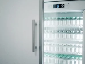 Farmaceutische koelkast op een kier met geordende medicijnflacons op witte planken en digitaal temperatuurscherm.