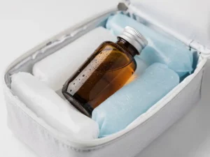 Amber glazen medicijnflesje in geïsoleerde koeltas met ijspakkingen op wit klinisch oppervlak, condensatie zichtbaar op het glas.